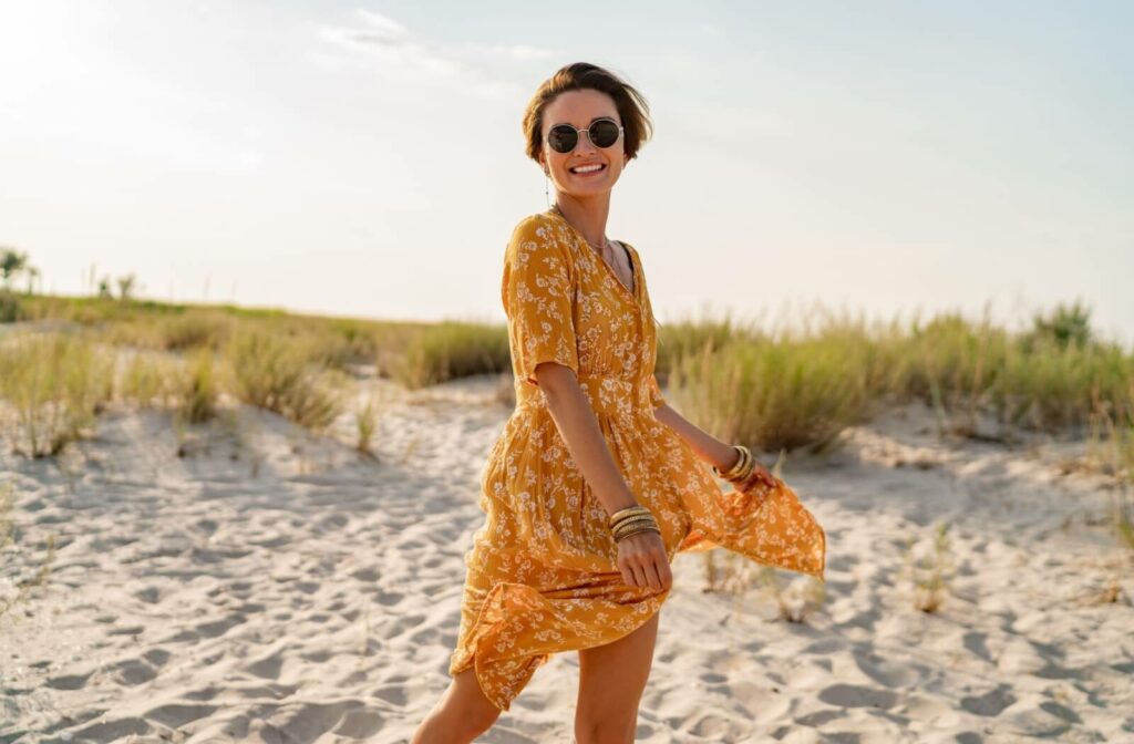 A stylish adult spins on a white, sandy beach while smiling. They are wearing a dark pair of polarized sunglasses.