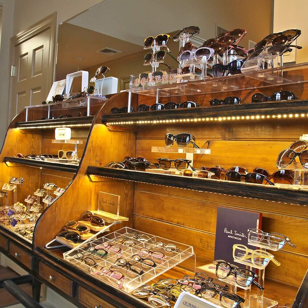 Display of luxury eyeglass and sunglass frames on wooden shelves in an Weston Optical, featuring designer brands like Oliver Peoples and Paul Smith under warm lighting.