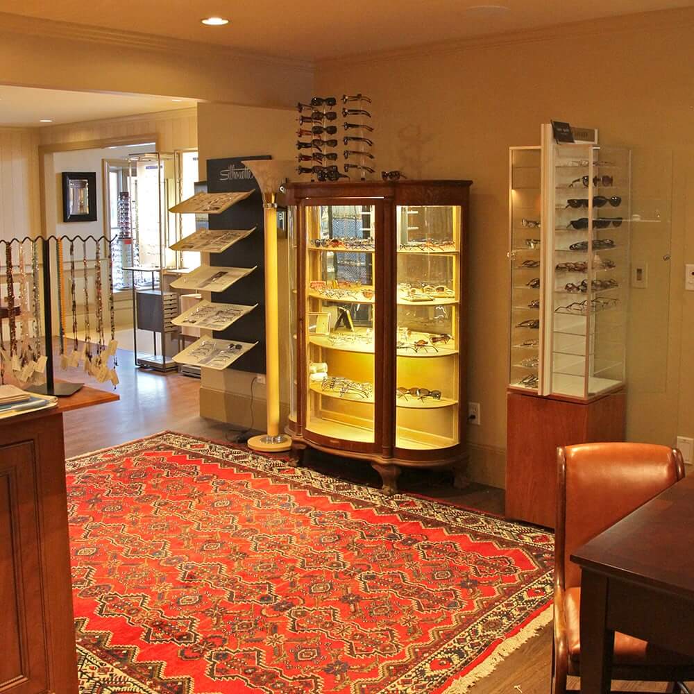 Elegant optical boutique interior featuring illuminated glass display cases filled with designer eyeglass frames, a vintage red patterned rug, and warm ambient lighting creating an upscale, inviting atmosphere.