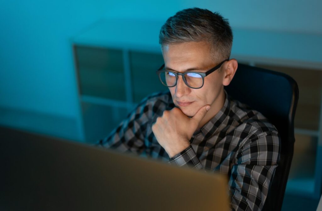 An individual wearing computer glasses with visible blue light reflections on the lenses while working at a monitor in a dim room.