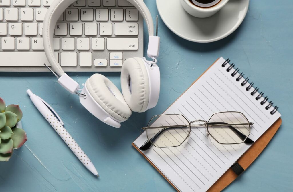 A flat-lay view of a blue desk featuring hexagonal computer glasses resting on a notepad next to white headphones and a keyboard.