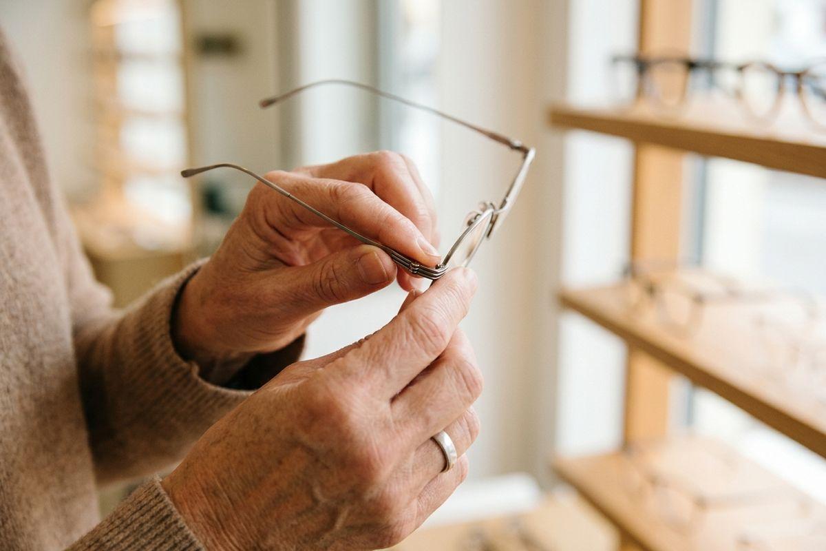 Person's hands holding thin-framed eyeglasses in an optician's shop, with more glasses on wooden shelves nearby.